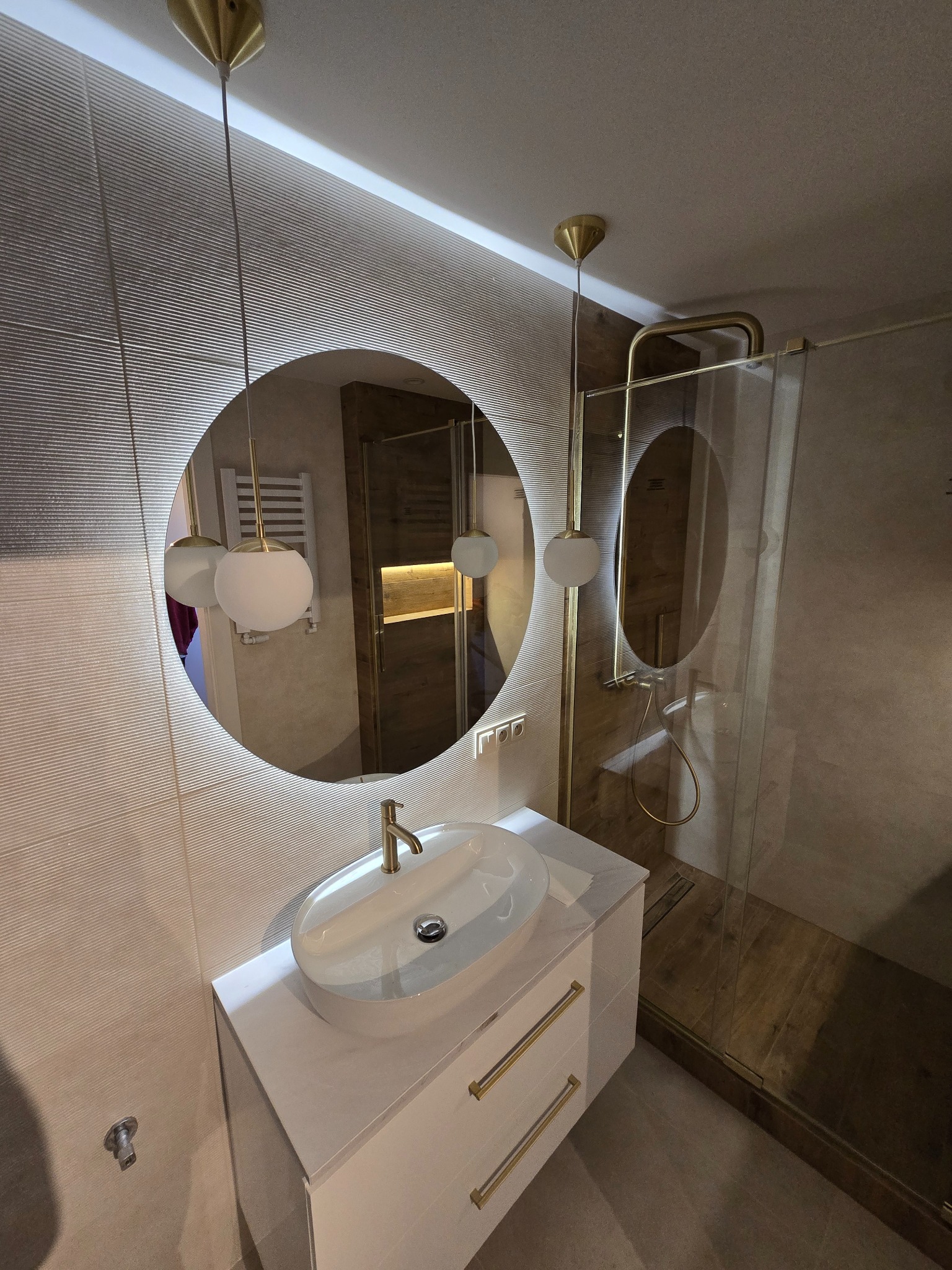 Wykończona łazienka z okrągłym lustrem z podświetleniem LED, białą umywalką nablatową na szafce z złotymi uchwytami i kabiną prysznicową ze złotymi ramami.