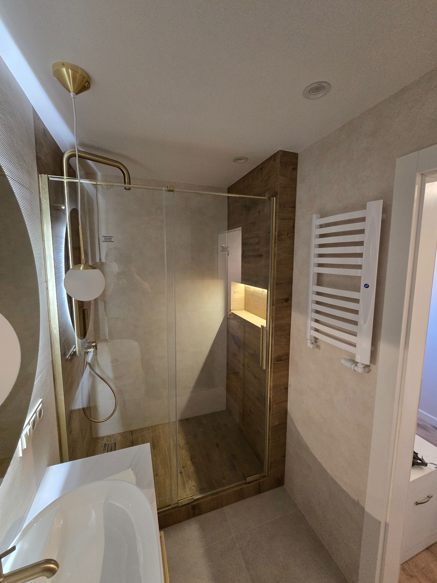 Nowoczesna łazienka po remoncie: przeszklona kabina prysznicowa z wnęką, grzejnik drabinkowy, umywalka z baterią w kolorze złota, oświetlenie punktowe.