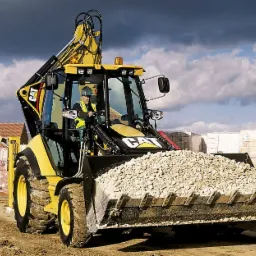 Żółta koparko-ładowarka marki CAT z łyżką wypełnioną jasnym kruszywem na placu budowy, w tle cegły i budynki mieszkalne pod pochmurnym niebem.
