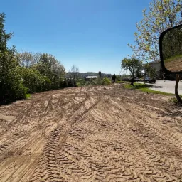 Widok z kabiny maszyny budowlanej na wyrównany teren z wyraźnymi śladami opon, w tle zabudowania i błękitne niebo.