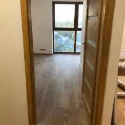Widok na pokój z nową podłogą laminowaną i drewnianą ościeżnicą drzwiową z przeszkleniami, widok z perspektywy korytarza, ściany pomalowane na biało, okno z widokiem na miasto w tle.