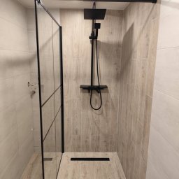 Patryk Kałkowski PatroBUDN1 - Nowoczesna kabina prysznicowa walk-in z czarnymi elementami, wykończona płytkami imitującymi drewno. Minimalistyczny design z odpływem liniowym.