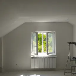 Poddasze po malowaniu z otwartym oknem, przez które widać zieleń drzew, grzejnik pod oknem, drabina z narzędziami malarskimi z boku.