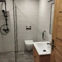 Nowoczesna łazienka z kabiną prysznicową typu walk-in z czarną armaturą, wiszącą toaletą i drewnianą szafką pod umywalką z czarnymi uchwytami.