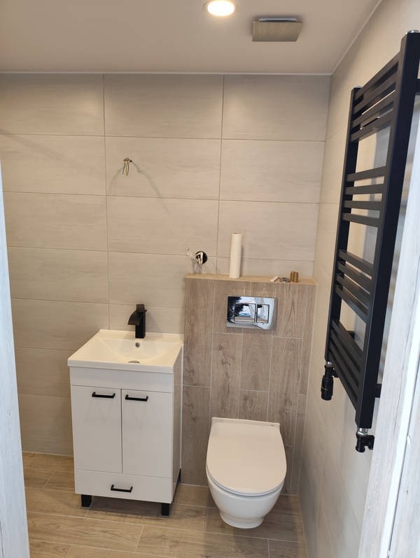 Nowoczesna łazienka z beżowymi płytkami, białą szafką pod umywalką z czarną armaturą i grzejnikiem drabinkowym. Uwagę zwraca drewnopodobna zabudowa toalety.
