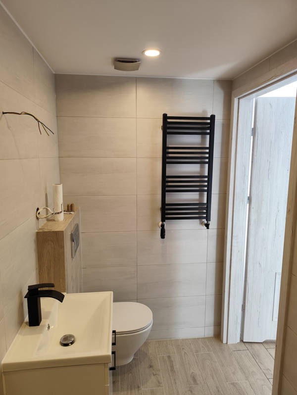 Jasna łazienka z beżowymi płytkami, czarną armaturą i grzejnikiem drabinkowym. Umywalka z szafką, toaleta i drewnopodobna podłoga tworzą spójną całość.