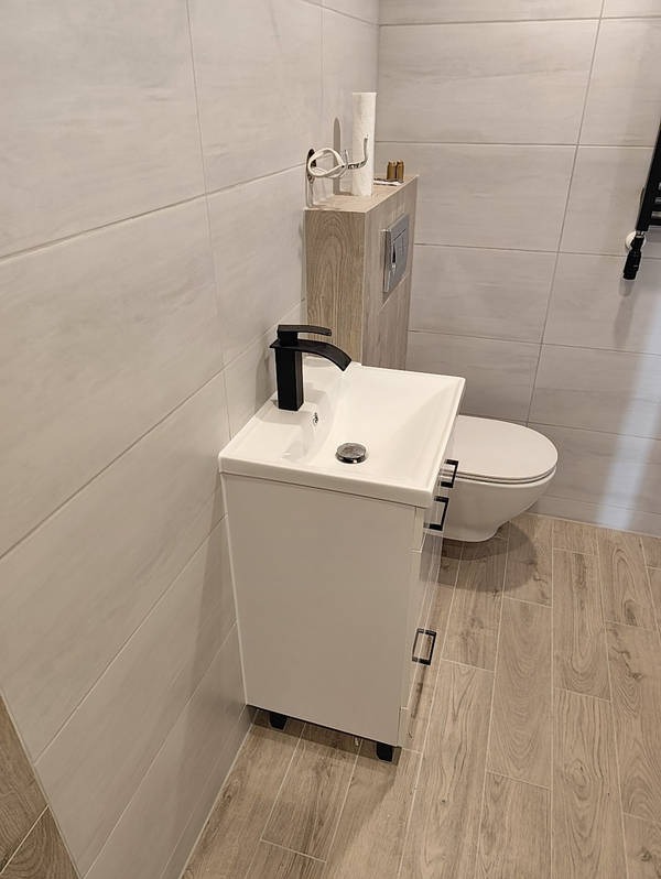 Nowoczesna łazienka z białą szafką pod umywalką i czarną armaturą, wykończona płytkami imitującymi drewno. Minimalistyczny design, jasne wnętrze.