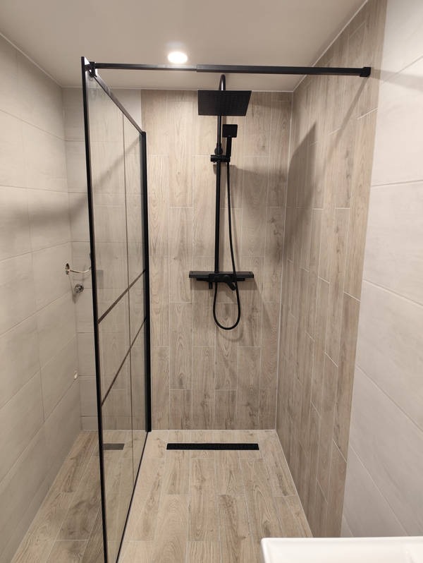 Nowoczesna kabina prysznicowa walk-in z czarnymi elementami, wykończona płytkami imitującymi drewno. Minimalistyczny design z odpływem liniowym.