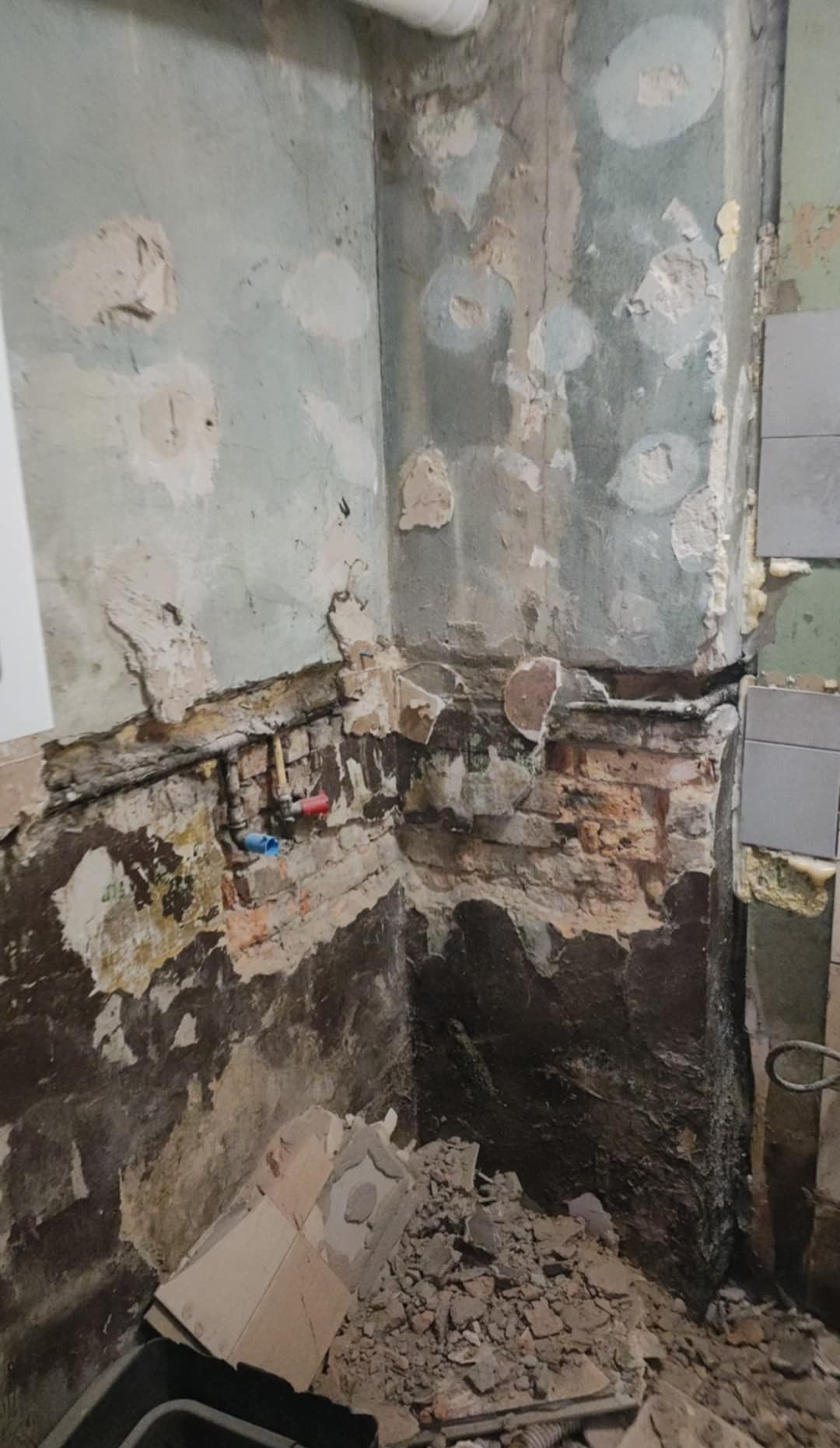 Odkryte instalacje wodne i fragmenty cegieł w rogu łazienki podczas remontu. Widoczne ślady po starych płytkach i resztki gruzu na podłodze.