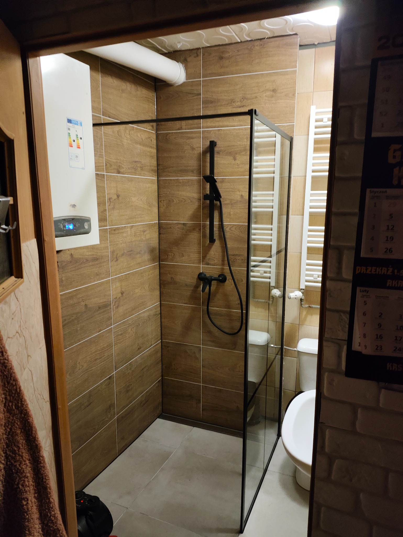 Wykończona łazienka z kabiną prysznicową typu walk-in z czarnymi profilami, brązowe płytki imitujące drewno, biały grzejnik drabinkowy i piec gazowy.