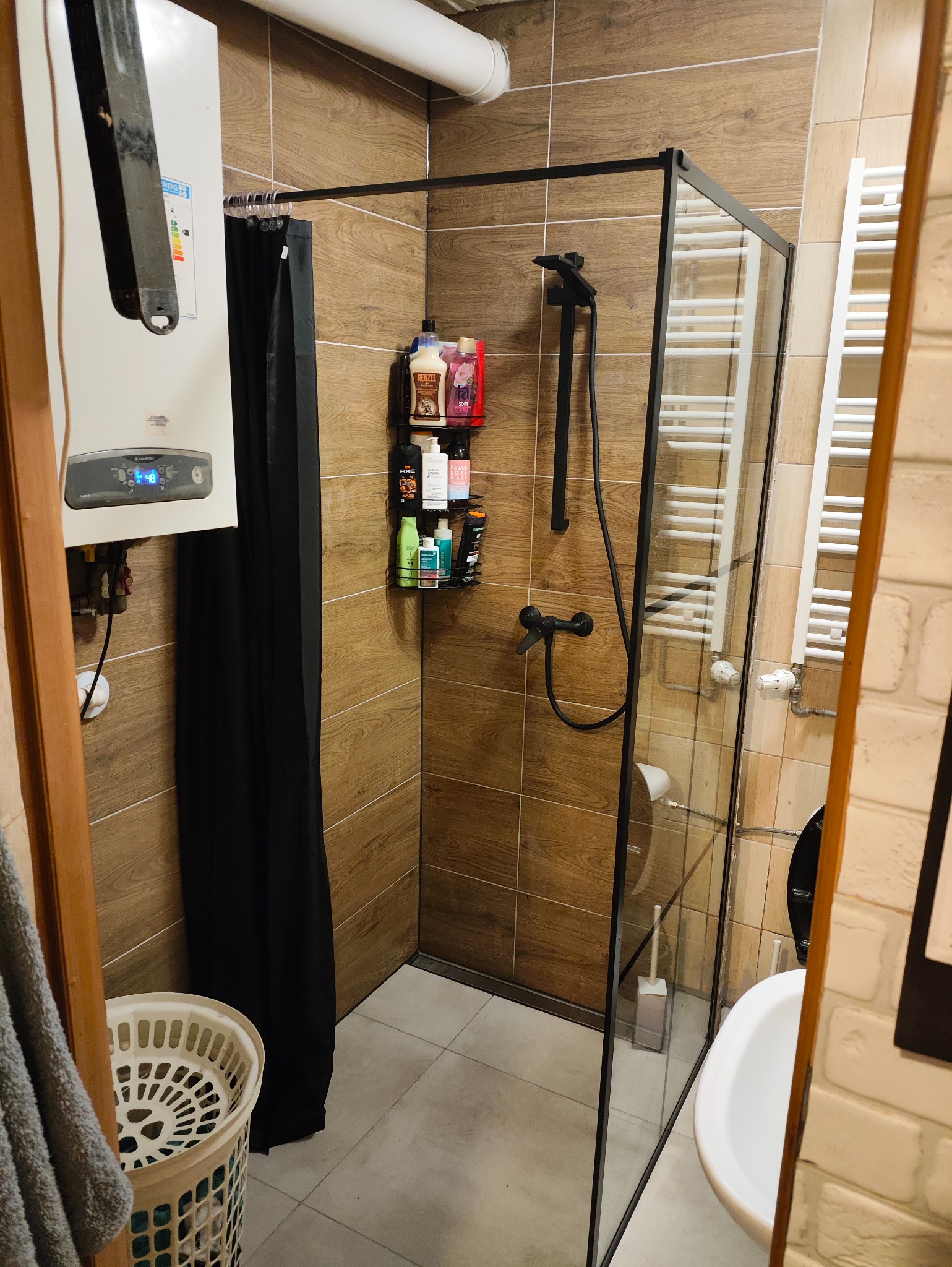 Nowoczesna kabina prysznicowa z czarną ramą i deszczownicą, drewnopodobne płytki na ścianach, wiszący grzejnik drabinkowy i bojler. Minimalistyczny design łazienki.