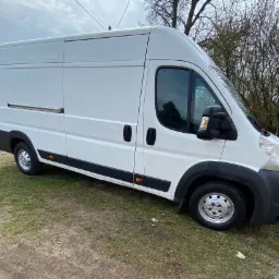 Biały samochód dostawczy z przesuwanymi drzwiami bocznymi, zaparkowany na trawiastym terenie w pochmurny dzień.