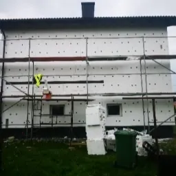 Dom jednorodzinny w trakcie ocieplania ścian zewnętrznych styropianem, widoczne rusztowania, fragmenty niezabezpieczonych otworów okiennych oraz składowany materiał izolacyjny.