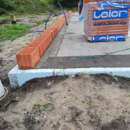 Budowa fundamentów: pierwszy rząd cegieł ceramicznych Leier ułożony na izolacji termicznej, widoczne niebieskie rury instalacyjne i paleta cegieł w tle.