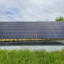 Duża instalacja paneli słonecznych na metalowej konstrukcji, widok z poziomu trawy, tło: błękitne niebo z chmurami.