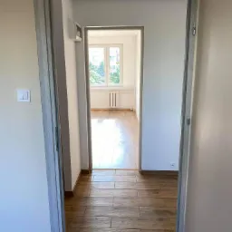 Widok przez odnowione drzwi z szarą ościeżnicą na drewnopodobną podłogę w wyremontowanym, jasnym pokoju z oknem i grzejnikiem.