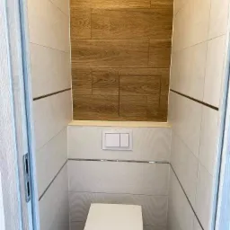 Nowoczesna toaleta w beżowo-brązowych płytkach z drewnopodobną wstawką, wisząca muszla klozetowa, ukryta spłuczka, wąskie pomieszczenie.