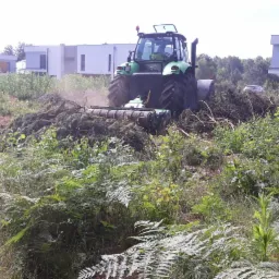 Zielony traktor z mulczerem leśnym rozdrabnia gałęzie i krzewy na zarośniętym terenie budowy, w tle nowoczesne domy.