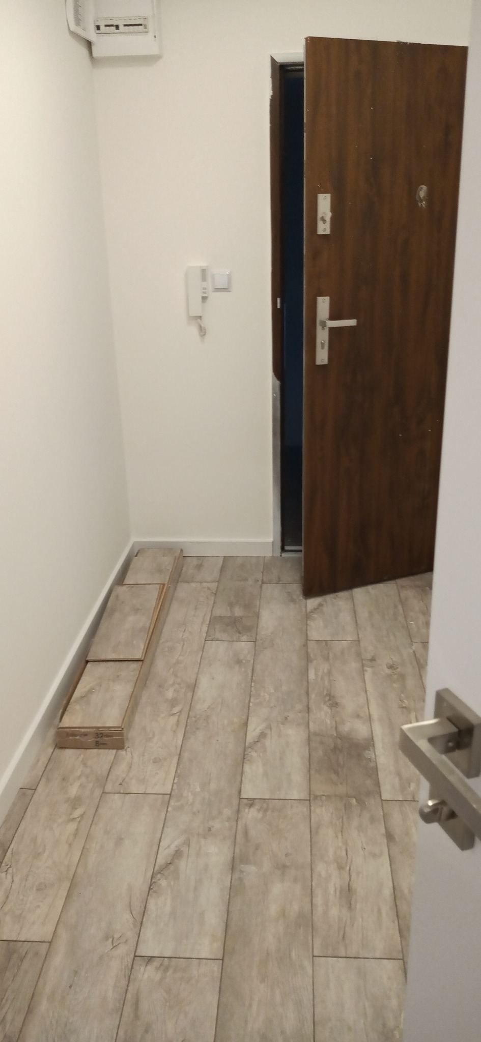 Korytarz po remoncie z nową podłogą z paneli imitujących drewno; paczka paneli leży przy ścianie, drzwi wejściowe są otwarte.