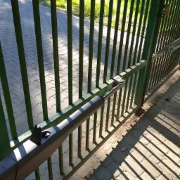 Zielona metalowa brama z zamontowanym siłownikiem automatycznym, rzucająca cienie na brukowaną nawierzchnię.