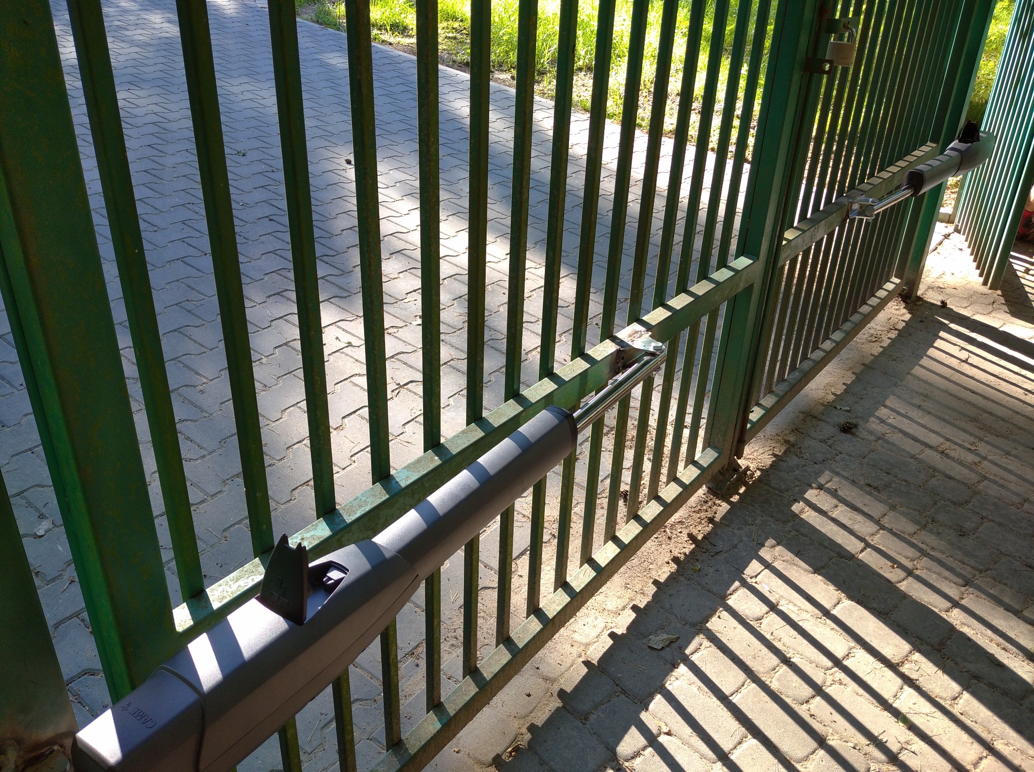 Zielona metalowa brama z zamontowanym siłownikiem automatycznym, rzucająca cienie na brukowaną nawierzchnię.
