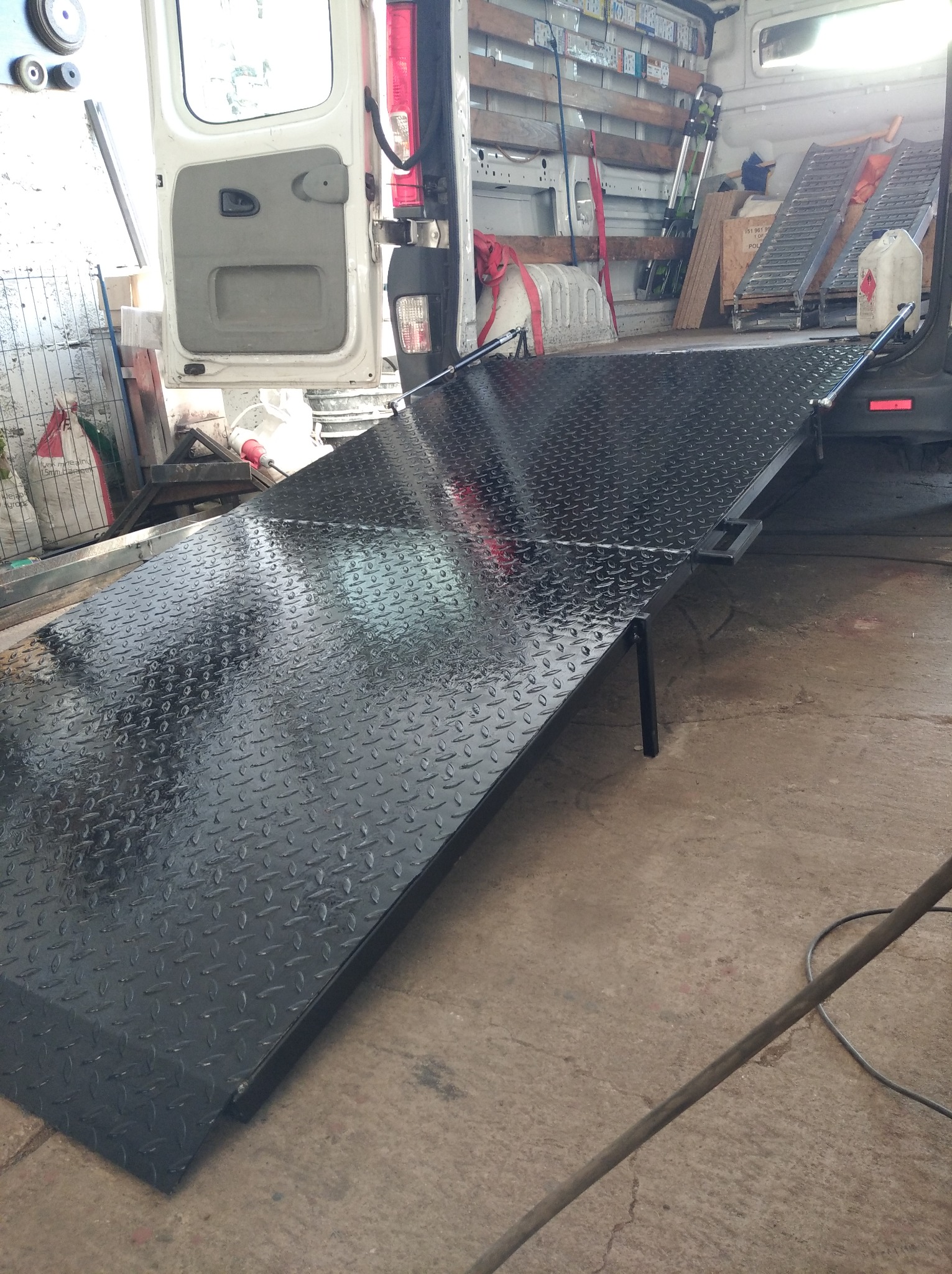 Czarna, metalowa rampa z antypoślizgową fakturą, wysunięta z otwartego bagażnika białego samochodu dostawczego z widocznymi narzędziami w środku.