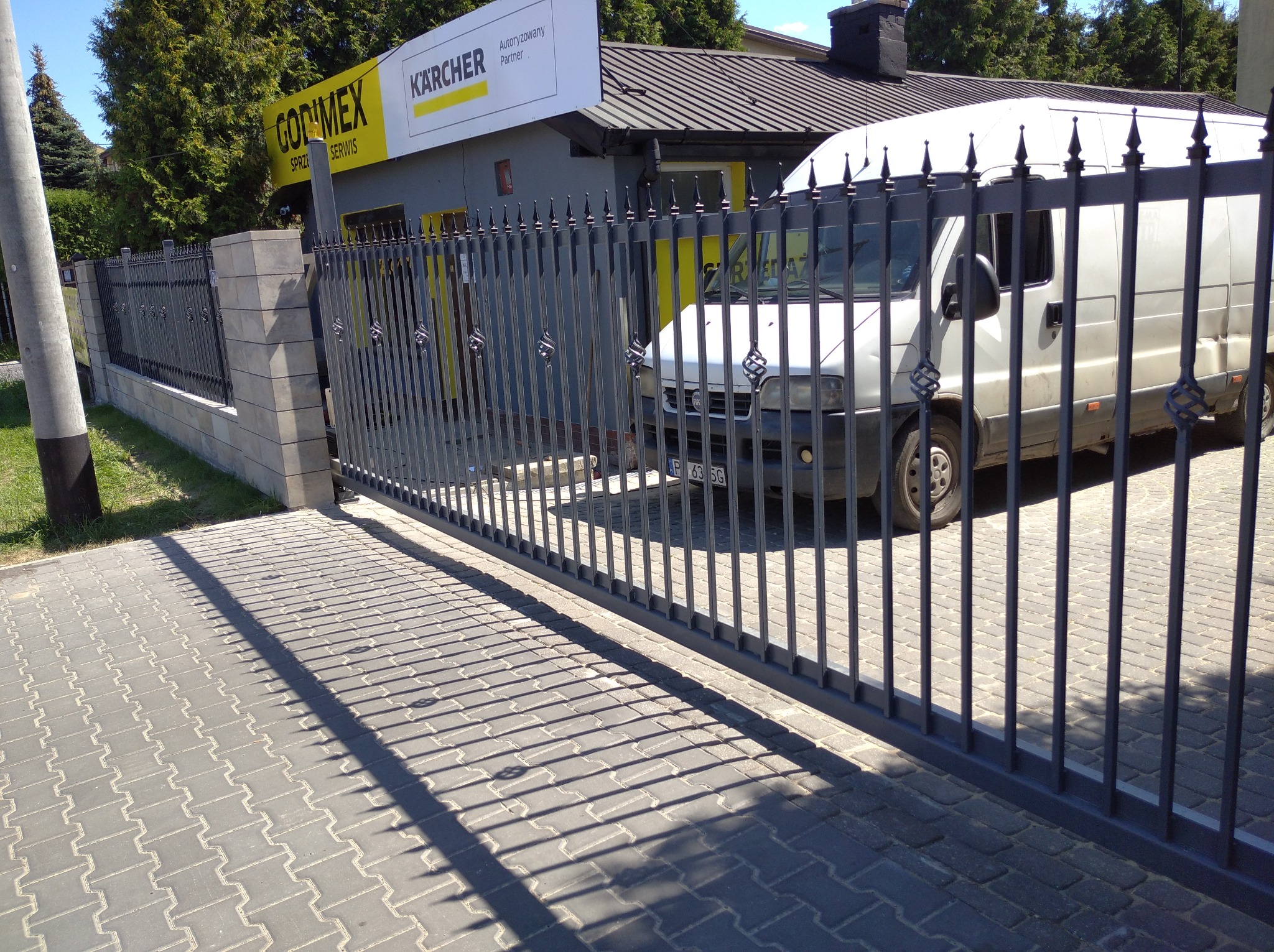 Przesuwna brama metalowa z ozdobnymi elementami w kształcie spirali, na tle budynku z szyldami firmowymi i zaparkowanym białym vanem.