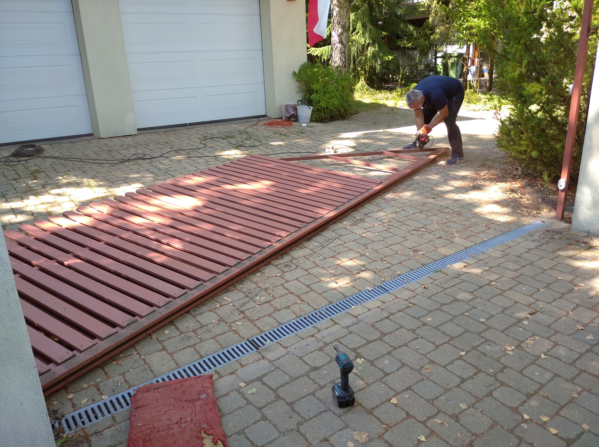 Pracownik w rękawicach tnie metalową ramę bramy na posesji z użyciem szlifierki kątowej, obok leży gotowe wypełnienie bramy z drewnianych listew.