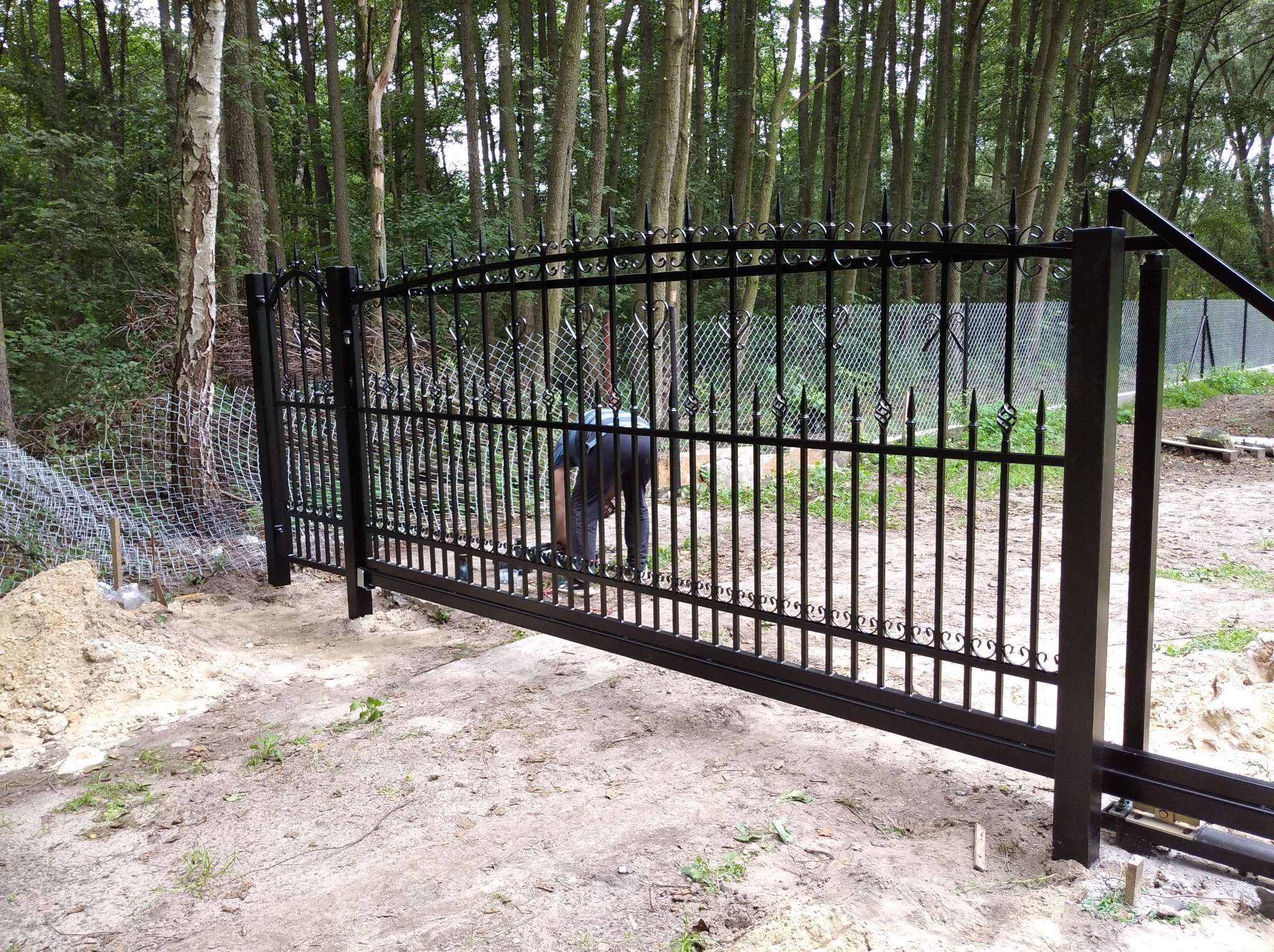 Montaż przesuwnej bramy z czarnego metalu z ozdobnymi detalami na tle leśnym, widoczna osoba w trakcie pracy.