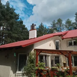 Drewniana konstrukcja tarasu przy domu z czerwonym dachem we Wrześni, z widocznymi roślinami pnącymi i elementami dekoracyjnymi, na tle drzew i błękitnego nieba.