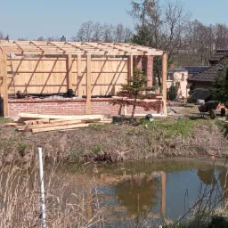 Konstrukcja drewniana na fundamencie z czerwonej cegły w trakcie budowy, widoczna z perspektywy stawu, z rozłożonymi deskami i drabiną opartą o ścianę.