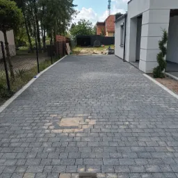 Szeroki podjazd z szarej kostki brukowej ułożonej we wzór jodełki, obrzeża z jasnych kostek, widoczne fragmenty ogrodzenia panelowego i bramy w kolorze grafitowym, w tle budynek mieszkalny...