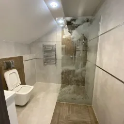 Wykończona łazienka na poddaszu ze szklaną kabiną prysznicową z motywem latarni morskiej, wiszącą toaletą i grzejnikiem drabinkowym.