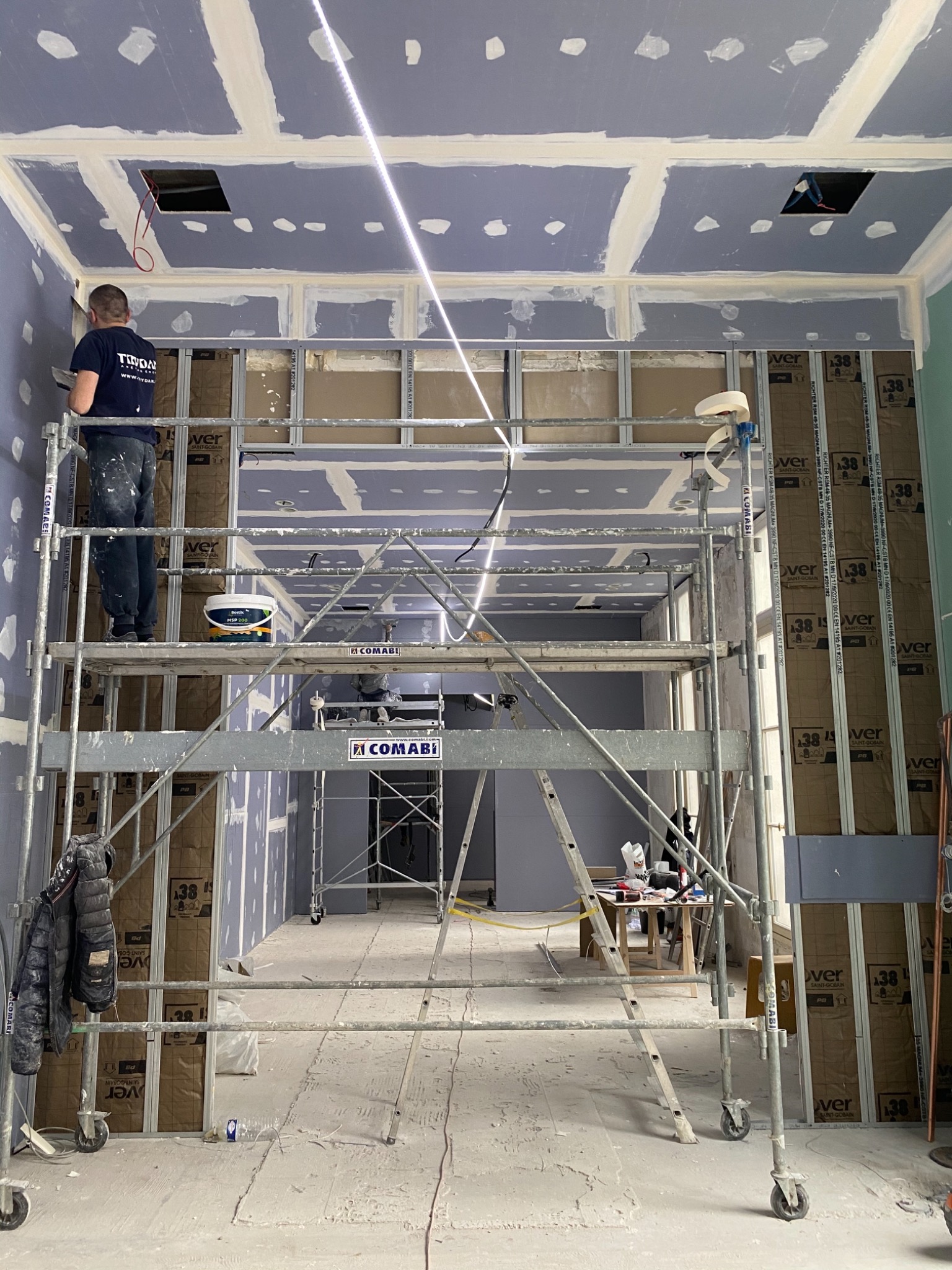 Pracownik na rusztowaniu szpachluje sufit podwieszany z widocznymi otworami na oświetlenie, ściany w trakcie wykańczania płytami gipsowo-kartonowymi, widoczne metalowe profile konstrukcyjne.