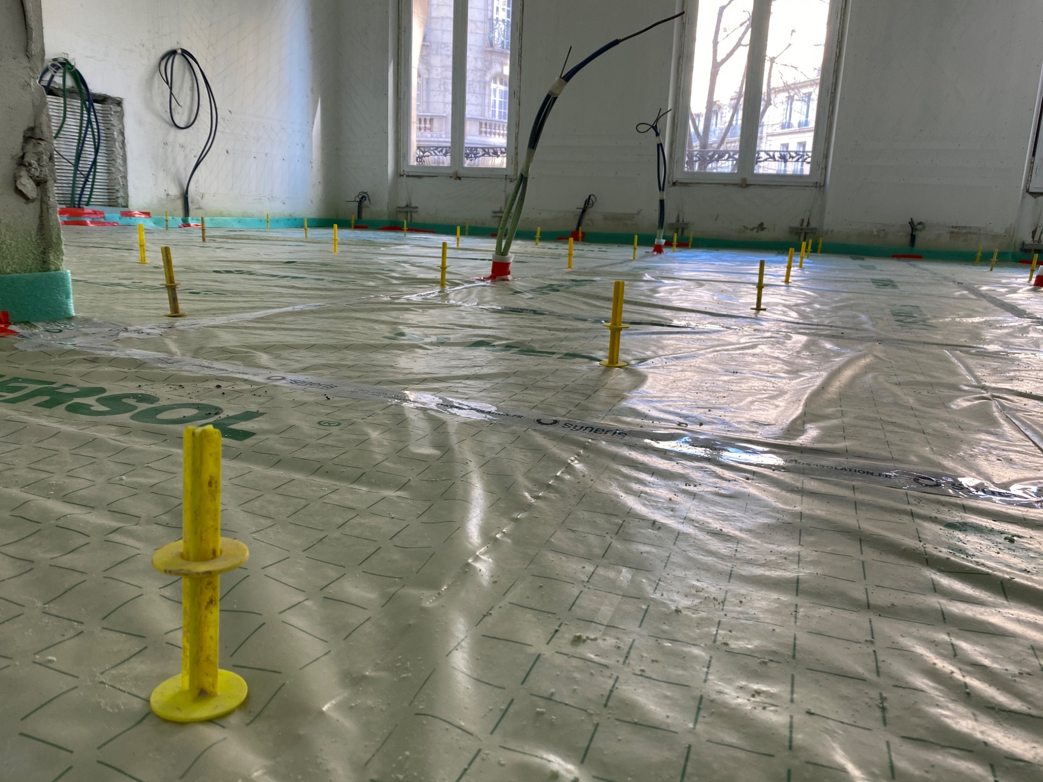 Przygotowanie podłoża pod ogrzewanie podłogowe: folia izolacyjna z nadrukiem kratki i żółte kołki poziomujące w surowym wnętrzu z oknami.