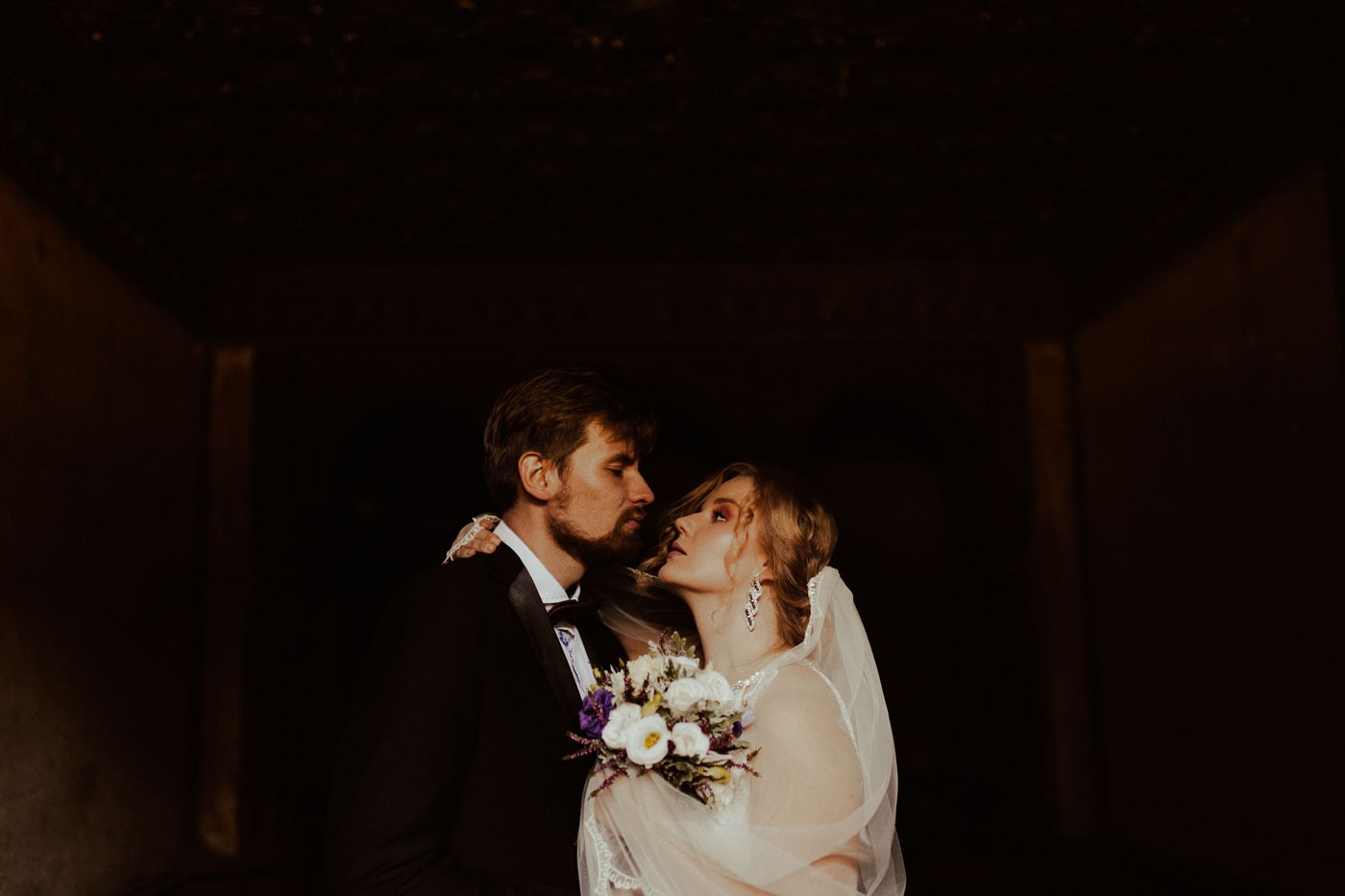 Para młoda wpatrzona w siebie w ciemnym przejściu, bukiet ślubny w dłoniach panny młodej, delikatna koronka na welonie.