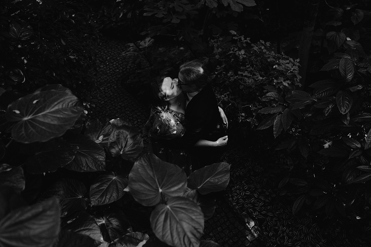 Czarno-białe zdjęcie pary młodej całującej się, otoczonej bujną roślinnością, z góry, z widoczną kamienną ścieżką w tle.