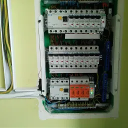 Otwarta skrzynka elektryczna z zabezpieczeniami różnicowoprądowymi i przepięciowymi, widoczne kable w listwach przypodłogowych na tle jasnozielonej ściany.