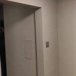 Cały Remontuje - Wykończone wnętrze z dekoracyjną fakturą na ścianach w odcieniach szarości, widoczny fragment podłogi z paneli, włącznik światła na ścianie oraz nowoczesna lampa sufitowa.