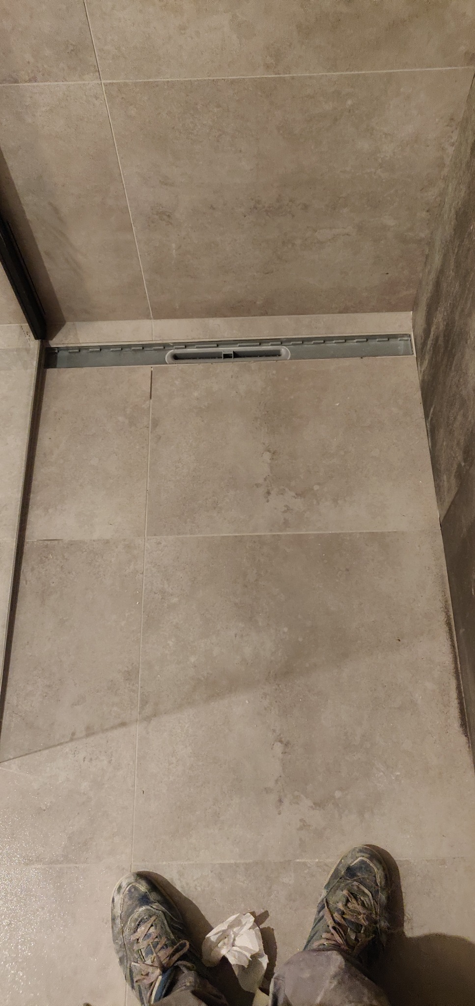 Beżowe płytki podłogowe w łazience z zamontowanym odpływem liniowym, widoczne robocze buty osoby wykonującej prace.