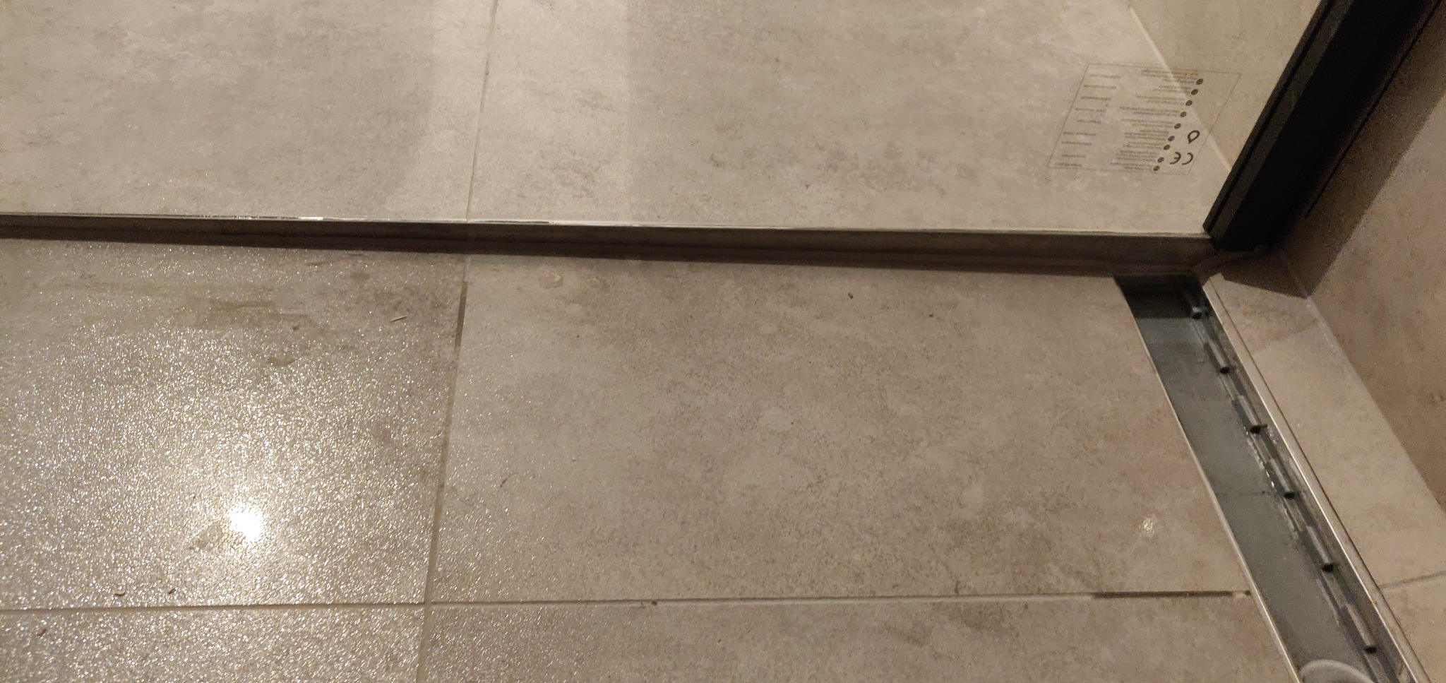 Szczegół odpływu liniowego w kabinie prysznicowej wyłożonej beżowymi płytkami, widoczna czarna rama szklanej ścianki i fragment naklejki informacyjnej na szkle.