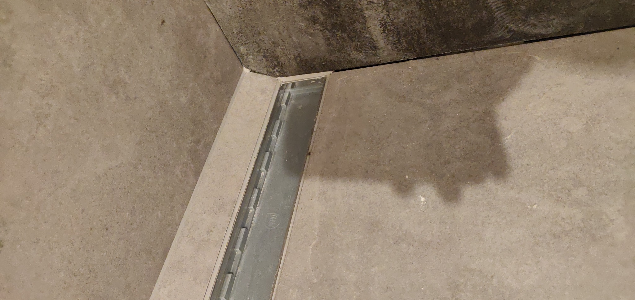 Ułożone płytki podłogowe w łazience z odpływem liniowym, widoczne fugi i szary sufit w rogu.