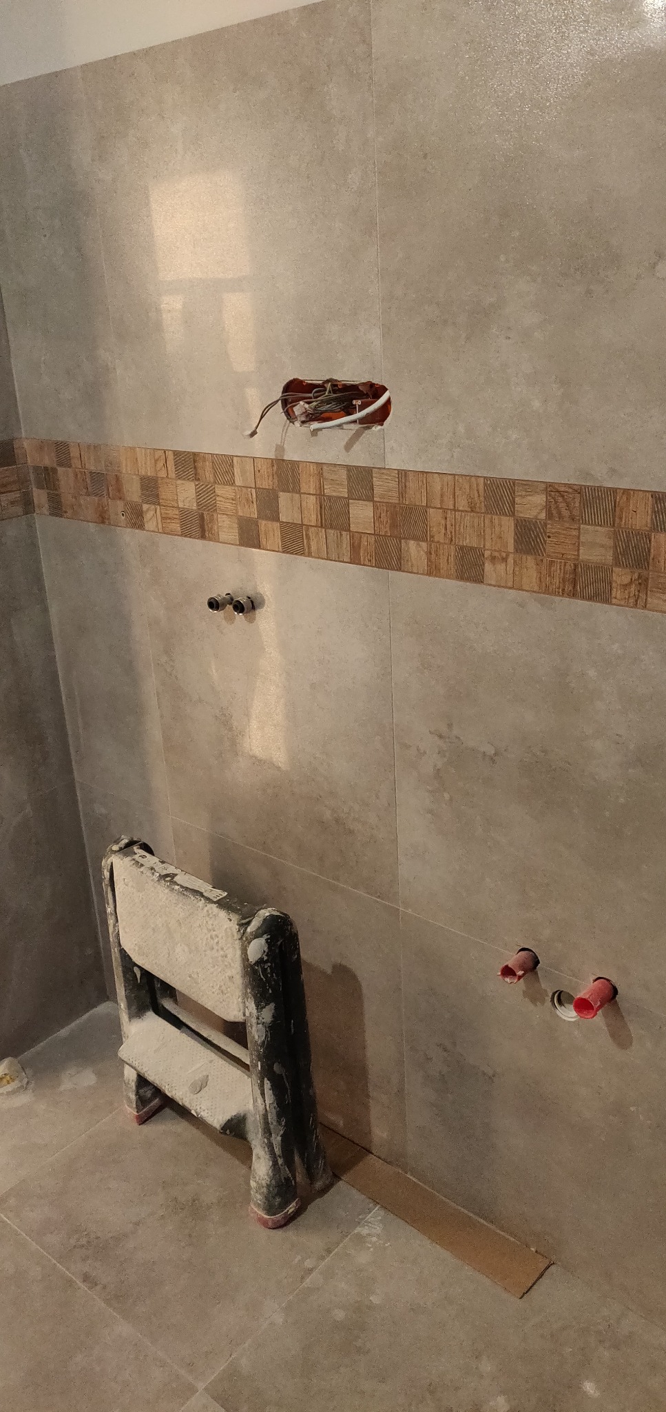 Ściana w trakcie wykańczania płytkami, widoczne otwory na instalacje, dekoracyjny pas płytek, drabina malarska pobrudzona zaprawą.