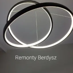 Nowoczesna lampa sufitowa z trzema okręgami LED o czarnych obudowach, zawieszona na cienkich linkach na tle białego sufitu.