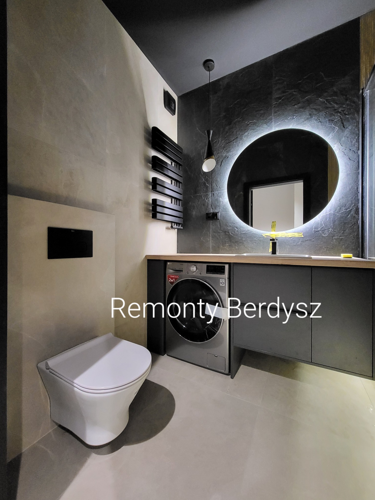 Nowoczesna łazienka z szarymi płytkami imitującymi kamień, podwieszaną toaletą, szafką z pralką i okrągłym lustrem z podświetleniem LED.