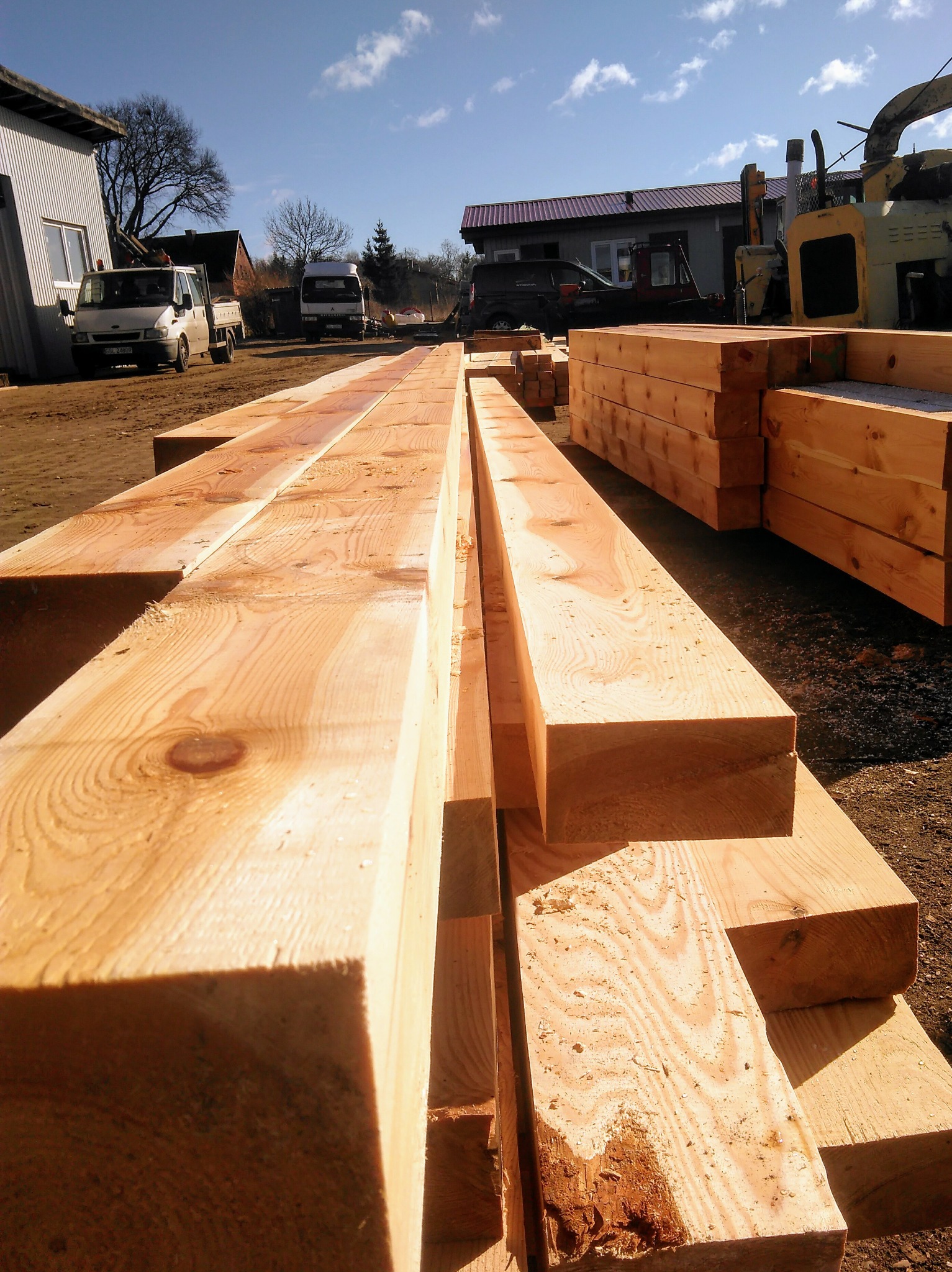 Stos jasnej tarcicy budowlanej ułożonej w długi rząd na placu składowym, w tle budynki gospodarcze i pojazdy.