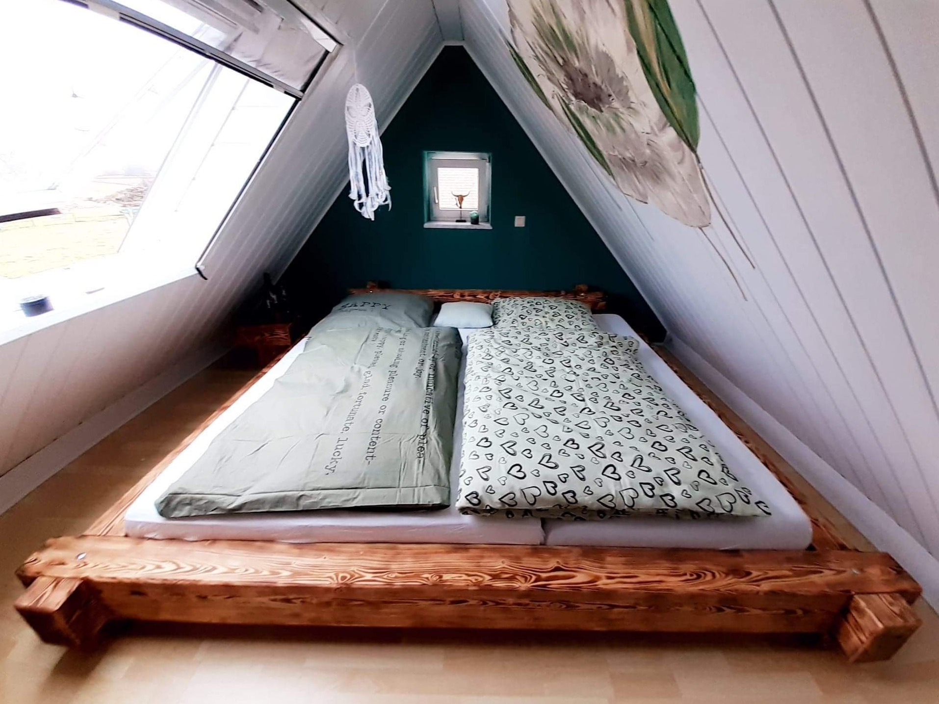 Drewniane łóżko w pokoju na poddaszu o skośnych ścianach, z oknem dachowym i dekoracjami w stylu boho.