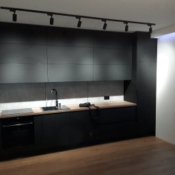 M.S. ALL-EURO Marcin Sobolewski - Nowoczesna kuchnia z matowymi, czarnymi frontami szafek, drewnianym blatem i oświetleniem punktowym na suficie. Na blacie czarny zlewozmywak i płyta indukcyjna. Na pierwszym planie parkiet...
