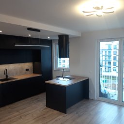 M.S. ALL-EURO Marcin Sobolewski - Nowoczesna kuchnia z czarnymi szafkami, drewnianym blatem i wyspą kuchenną z płytą indukcyjną, oświetlona lampą sufitową w kształcie kwiatu i podłużną lampą nad blatem.