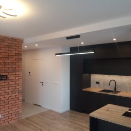 M.S. ALL-EURO Marcin Sobolewski - Nowoczesna kuchnia z czarnymi szafkami, drewnianymi blatami i ceglaną ścianą z uchwytem na telewizor, oświetlona designerskimi lampami sufitowymi.